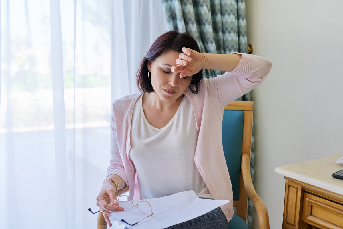 Stress, headache, migraine, upset middle-aged woman holding her head with her hands, sitting at home on a chair with papers in her hands. Health problems, age-related changes, mature women